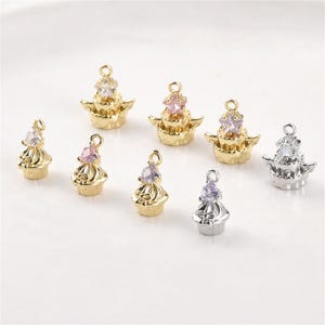 Peut inclure: Une collection de petits pendentifs en métal en forme de cupcakes et de gâteaux à étages, avec des finitions dorées et argentées. Chaque breloque est surmontée d'un cristal facetté transparent, rose ou violet, et possède une petite boucle.