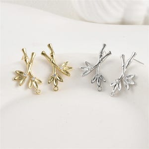 Op de afbeelding: Twee paar oorbellen in goud- en zilverkleur. Elke oorbel heeft een bamboestengelontwerp met drie bladeren en een kleine, heldere edelsteen. De oorbellen hebben een lus aan de onderkant om aan andere sieraden te bevestigen.