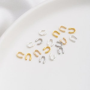 以下が含まれることがあります： 銀色と金色をした小さな金属製のジャンプリングの集まり。リングはU字型で、白い表面に散らばっています。