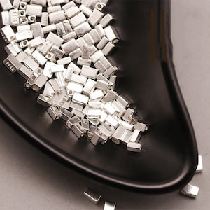 Puede incluir: Primer plano de un cuenco negro lleno de cuentas espaciadoras de metal plateado. Las cuentas son de forma rectangular con un pequeño agujero en el centro.