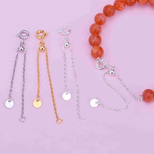 Puede incluir: Tres cadenas de extensión de pulsera ajustables en plata y oro con un pequeño disco redondo al final de cada cadena. Las cadenas están sobre un fondo rosa.