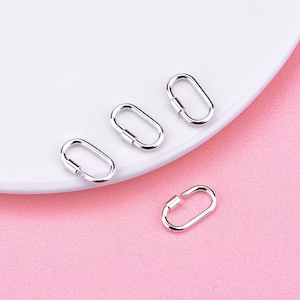May include: Four small, oval-shaped silver clasps with a screw-on closure are arranged on a white plate against a pink background. The clasps are shiny and reflect the light, suggesting a polished finish. The design is simple and modern.