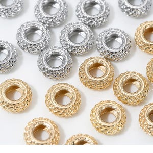 Może przedstawiać: Zbliżenie na wiele srebrnych i złotych metalowych koralików. Koraliki mają teksturowany, okrągły wzór z otworem pośrodku, odpowiednim do nawlekania. Koraliki są prawdopodobnie przeznaczone do tworzenia biżuterii lub innych projektów rzemieślniczych.