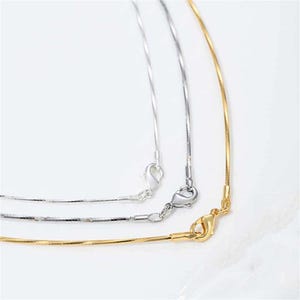 May include: Three delicate necklaces in silver and gold. The necklaces have a snake chain design and are displayed against a white background. Each necklace has a lobster clasp closure.