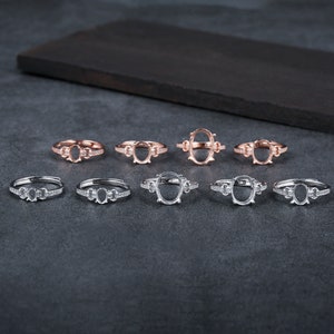 Puede incluir: Diez anillos de plata y oro rosa con engastes ovalados y pequeñas piedras transparentes alrededor de los engastes. Los anillos están dispuestos en dos filas sobre una superficie gris.
