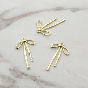 Op de afbeelding: Drie goudkleurige metalen bedels met een strikachtig ontwerp. Elke bedel heeft een lusvormige bovenkant en drie hangende, langwerpige staven. De bedels zijn gerangschikt op een lichtgekleurd, gemarmerd oppervlak.