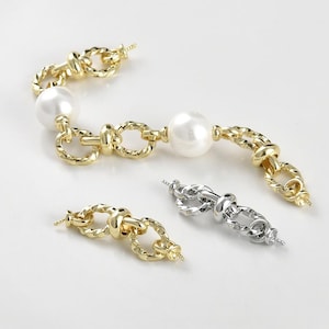 Puede incluir: Dos eslabones de cadena dorados y uno plateado con una perla en cada extremo. Los eslabones de cadena están retorcidos y tienen un diseño de nudo. Las perlas son blancas y redondas.