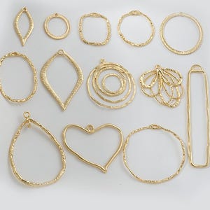 May include: A set of 13 gold-toned metal pendant blanks with various shapes, including circles, ovals, squares, hearts, and a teardrop. The blanks have a textured surface and are ready for crafting.