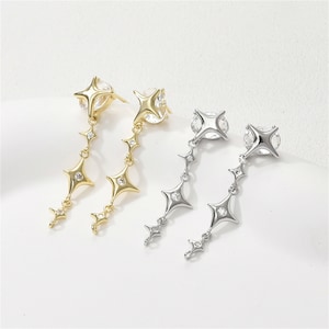 Pode incluir: Brincos pendurados em tons de ouro e prata. Cada brinco apresenta um pino em forma de estrela com uma pedra transparente grande. Uma corrente de pingentes em forma de estrela menores desce do pino, criando um design celestial delicado. Fundo branco.