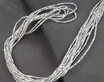 Puede incluir: Un collar de cadena de plata hecho de pequeñas cuentas facetadas. La cadena está en capas y drapeada sobre una superficie gris oscuro.