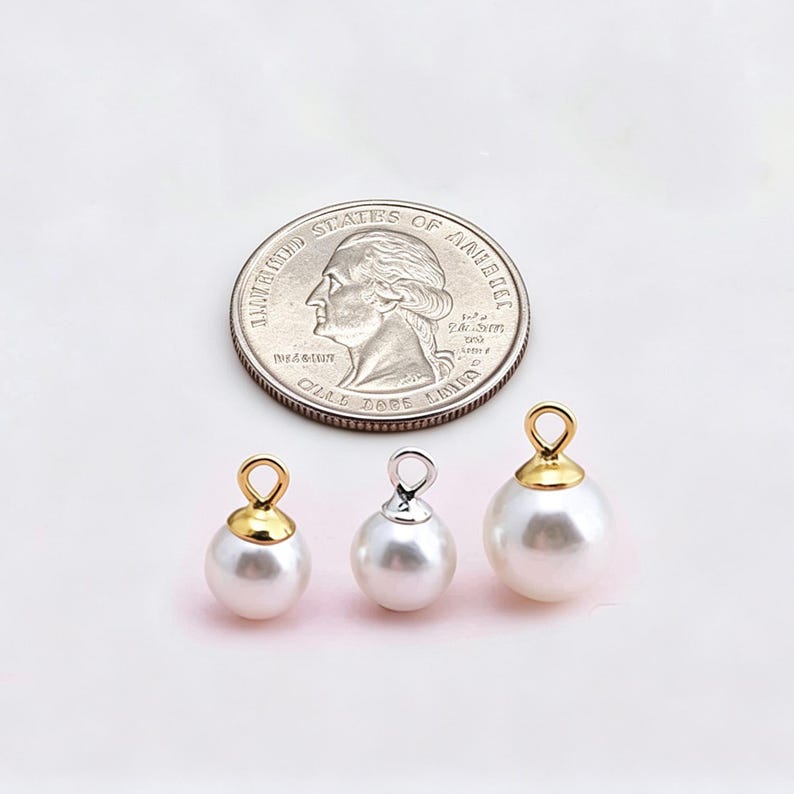 Puede incluir: Tres dijes de perlas blancas esf&eacute;ricas con anillas met&aacute;licas en la parte superior, colocados debajo de una moneda de cuarto de d&oacute;lar estadounidense para comparar el tama&ntilde;o. Dos dijes tienen tapas doradas y el central tiene tapa plateada.