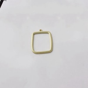 May include: A gold-colored, square pendant with rounded corners and a small loop at the top. The pendant is a simple, geometric shape, suitable for jewelry making or crafting. The pendant is shown against a white background.