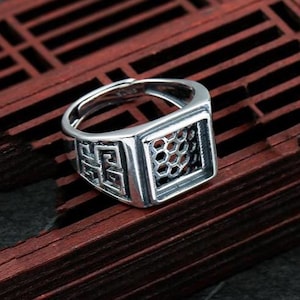 May include: A silver-colored ring with a square face featuring a honeycomb pattern and decorative side detailing. The ring is adjustable and sits on a dark wooden surface, showcasing its intricate design and craftsmanship.