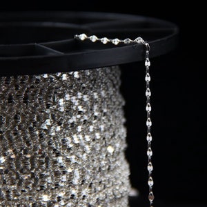 Puede incluir: Un carrete de cadena plateada con un solo hilo que cuelga. La cadena está hecha de pequeños eslabones redondos.