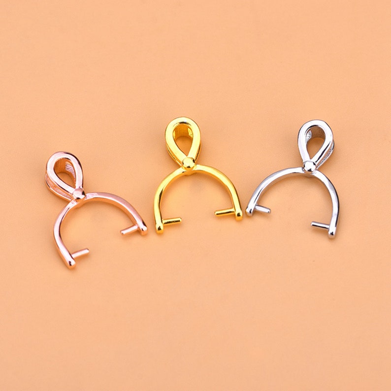 May include: Three metal pendant bails in rose gold, gold, and silver. Each bail has a curved base with two small posts and a loop at the top for attaching to a chain or cord. The bails are displayed against a peach background.
