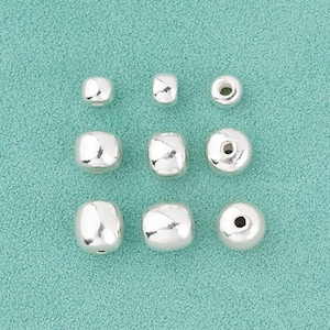 Peut inclure: Neuf perles argentées de formes variées, dont des cubes et des sphères, sont disposées sur un fond turquoise. Les perles ont une surface brillante et réfléchissante, destinées à la création de bijoux ou à l'artisanat.