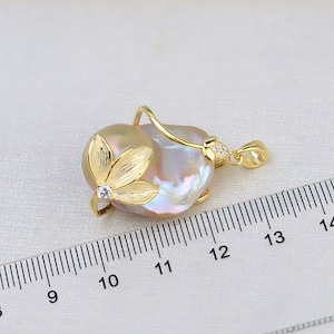 Op de afbeelding: Een gouden hanger met een grote, iriserende barokparel met een delicaat gouden bloemmotief. De parel is ongeveer 2,5 cm in diameter.