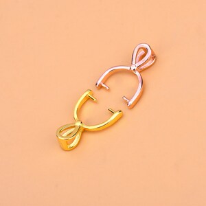 May include: Two pendant bails, one gold-colored and one rose gold-colored, are displayed against a peach background. The bails are open, showcasing their curved design with a loop at the top and two prongs extending from the bottom.