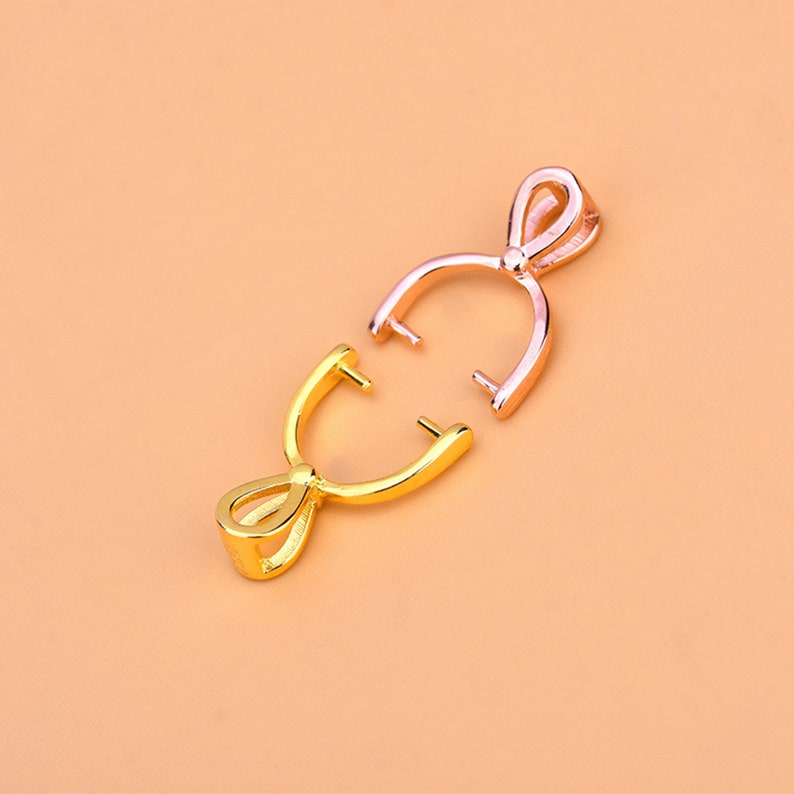 May include: Two pendant bails, one gold-colored and one rose gold-colored, are displayed against a peach background. The bails are open, showcasing their curved design with a loop at the top and two prongs extending from the bottom.