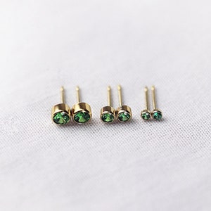 May include: Four pairs of gold-colored stud earrings with round, emerald-green gemstones. The earrings vary in size, with the largest pair on the left and the smallest on the right. The earrings are displayed on a white, textured surface.