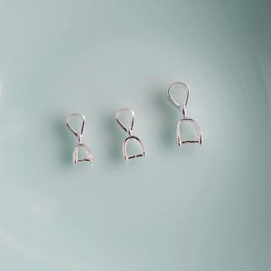 Puede incluir: Tres pequeños hallazgos de joyería plateados, cada uno con un lazo en la parte superior y una base en forma de D. Los hallazgos están dispuestos sobre una superficie azul claro, creando una estética minimalista. Estos hallazgos probablemente se utilizan en la fabricación de joyas.