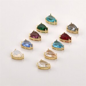 May include: A collection of ten teardrop-shaped jewelry charms, each featuring a faceted gemstone set in a gold-toned metal frame with a small loop at the top. The stones come in various colors including clear, white, blue, green, red, and purple.