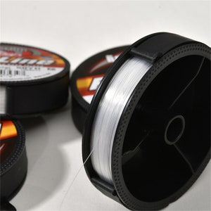 May include: Close-up of a spool of clear fishing line in a black plastic dispenser. The line is wound around a central core and the dispenser has a black lid with a small tab.