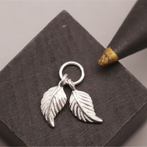 May include: A silver charm with two small leaf pendants hanging from a circular ring. The charm is on a dark gray surface.