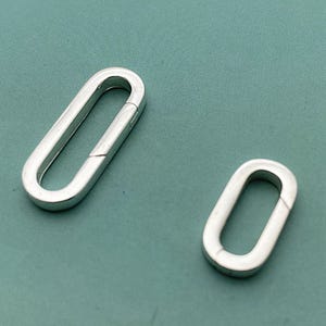 May include: Two silver-colored oval-shaped clasps. One clasp is open, revealing the hinge mechanism. The other clasp is closed. The clasps are on a teal-colored surface. These clasps are suitable for jewelry making and other craft projects.
