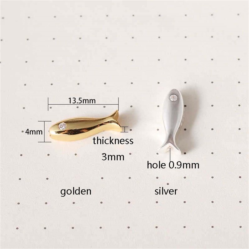 May include: Two small fish-shaped charms, one gold and one silver, with a clear rhinestone in the centre. The gold charm measures 13.5 mm long and 4 mm wide, with a 3 mm thickness. The silver charm has a 0.9 mm hole.