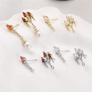 May include: A collection of gold and silver-toned earring components featuring trident-shaped studs with orange or clear teardrop-shaped stones. Some pieces include dangling white pearls or small loops for attachments, showcasing intricate metalwork designs.