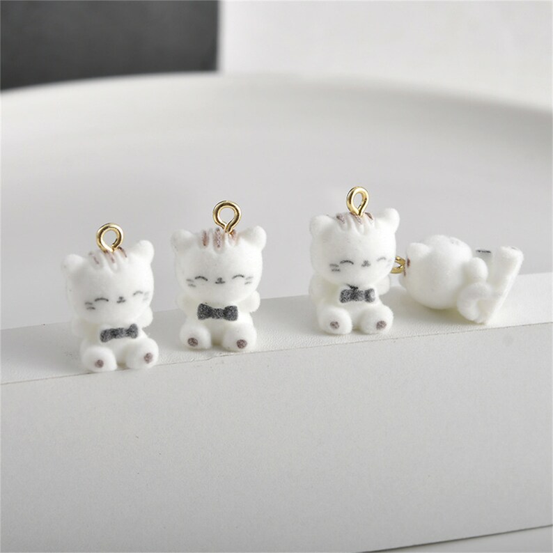 May include: Three white ceramic cat charms with gold jump rings. Each charm features a black bow tie and a brown stripe on its head. The cats are sitting with their paws together.