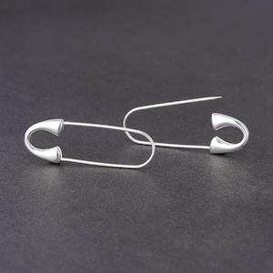 May include: A pair of silver-colored safety pin earrings. The earrings are in the shape of large safety pins, with a curved top and a pointed end. The earrings are displayed against a dark background.