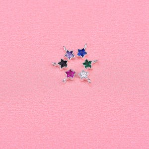 May include: Six silver star-shaped charms with different colored gemstones. The charms have a loop at the top for attaching to a necklace or bracelet. The gemstones are purple, blue, green, red, clear, and black.