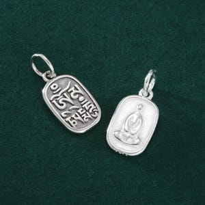 Puede incluir: Dos colgantes plateados con temática religiosa. Uno presenta una inscripción, el otro representa una figura sentada en meditación. Ambos tienen un lazo para una cadena y están sobre un fondo verde oscuro.