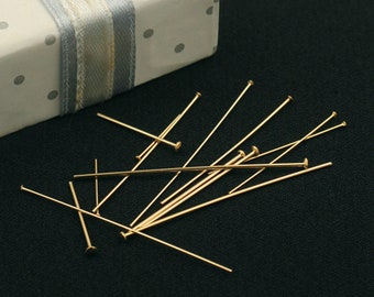 May include: A group of gold-colored head pins scattered on a black surface. The pins are thin and straight, with a small round head at one end.