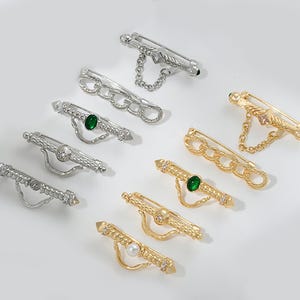 May include: A set of six gold and silver brooches with ornate designs. The brooches feature a variety of embellishments, including chains, green and white stones, and small beads.