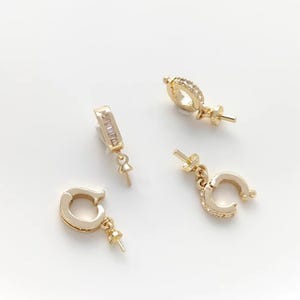 May include: Four gold-colored earring components with various designs. One is a rectangular bar with clear stones, and the others are curved or circular with small, clear stones. Each has a post for attaching to a backing.