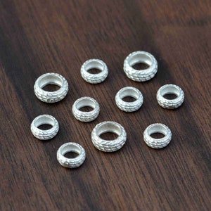 May include: A collection of small, silver-colored, textured metal beads. The beads are round with a central hole and a detailed, patterned surface. They are scattered on a dark brown wooden surface.