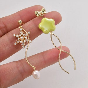 May include: A pair of gold dangle earrings. One earring features a snowflake with clear crystals and a pearl drop. The other earring features a yellow flower charm and a bow.