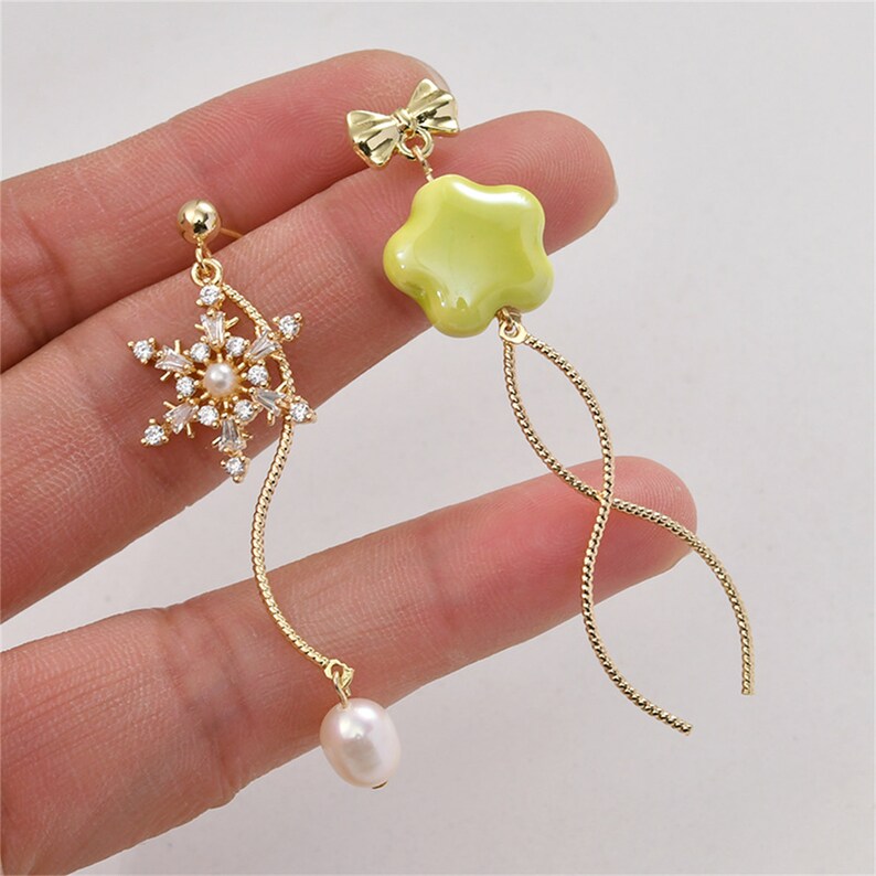 May include: A pair of gold dangle earrings. One earring features a snowflake with clear crystals and a pearl drop. The other earring features a yellow flower charm and a bow.