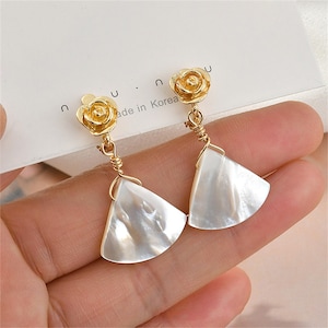 May include: A pair of gold earrings with a white shell-shaped pendant and a gold rose charm. The earrings are hanging from a person's ear.