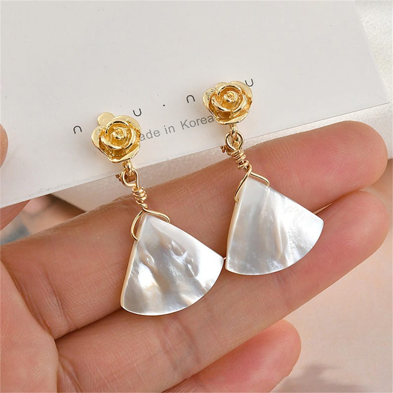 May include: A pair of gold earrings with a white shell-shaped pendant and a gold rose charm. The earrings are hanging from a person's ear.