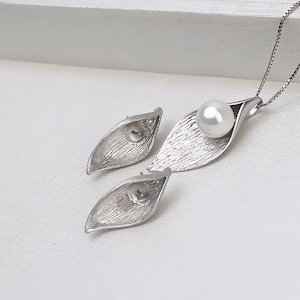 May include: A silver necklace and earrings set featuring a delicate leaf design with a single pearl pendant. The necklace has a silver chain and the earrings are stud style.