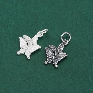 May include: Two silver butterfly charms with detailed wing patterns. Each charm has three butterflies, with a small loop for attaching to jewellery. The charms are set against a dark green backdrop.