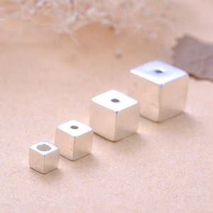 Puede incluir: Cuatro cuentas de cubo de plata de diferentes tamaños, dispuestas en una línea de la más pequeña a la más grande. Cada cubo tiene un pequeño agujero en el centro de un lado.