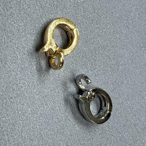 May include: Two metal jewelry clasps. One is gold-toned, the other is silver-toned. Both clasps have a circular design with a hook and loop closure. The clasps are on a gray surface.