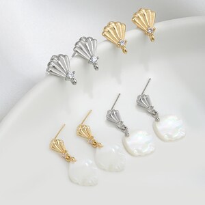 May include: A selection of earrings in gold and silver hues. Some earrings have a fan-shaped design with a small gemstone, while others feature a pearlescent, teardrop-shaped charm. The earrings are presented on a white surface.
