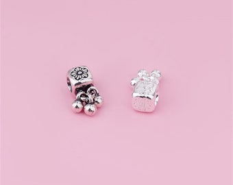 Könnte beinhalten: Zwei silberne Charms mit Blumenmotiven. Ein Charm hat einen Blumenanhänger mit drei baumelnden Glöckchen. Der andere Charm hat ein Blumenmuster.