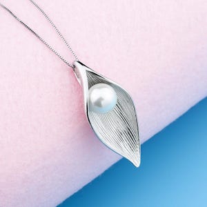 Può includere: Una collana d'argento con un ciondolo a forma di foglia, che tiene una perla bianca al centro.